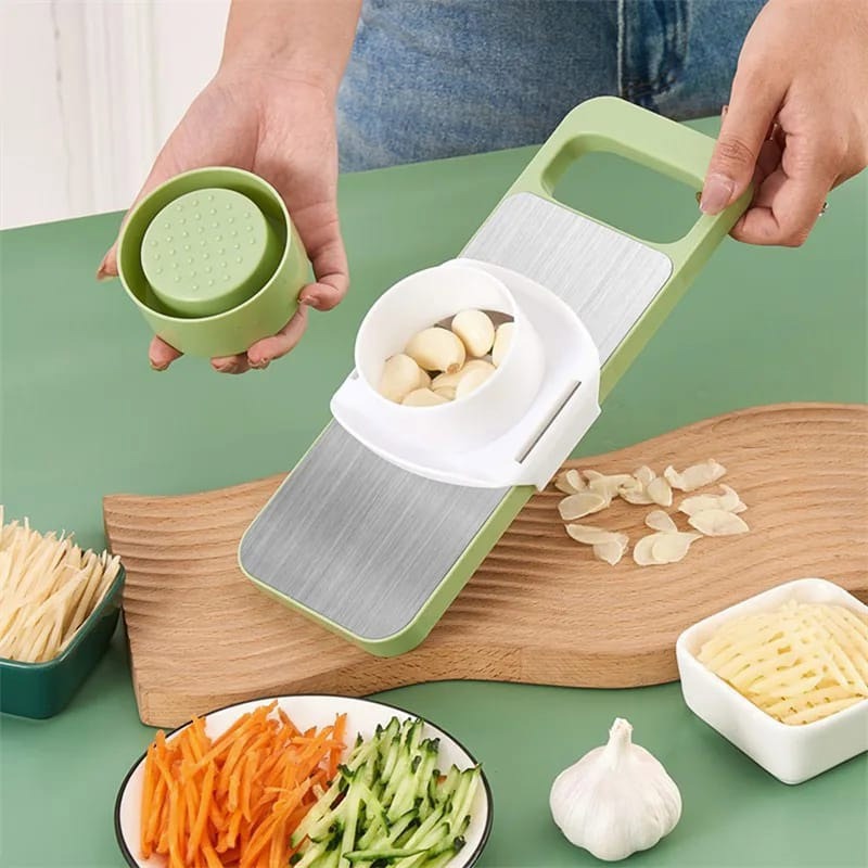 Green kitchen tool with garlic being sliced on a green table with vegetables and a bowl.